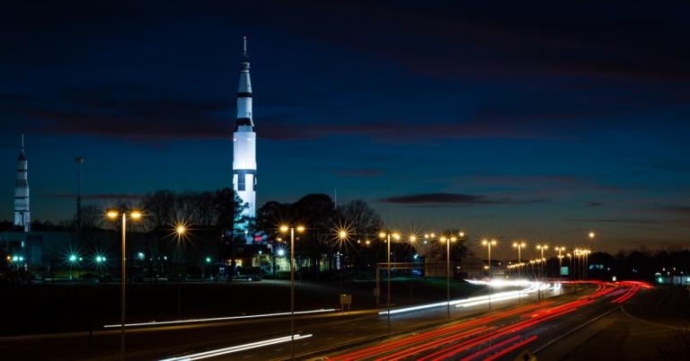 Rocket on its launch pad at dusk with light trails from traffic on a nearby highway